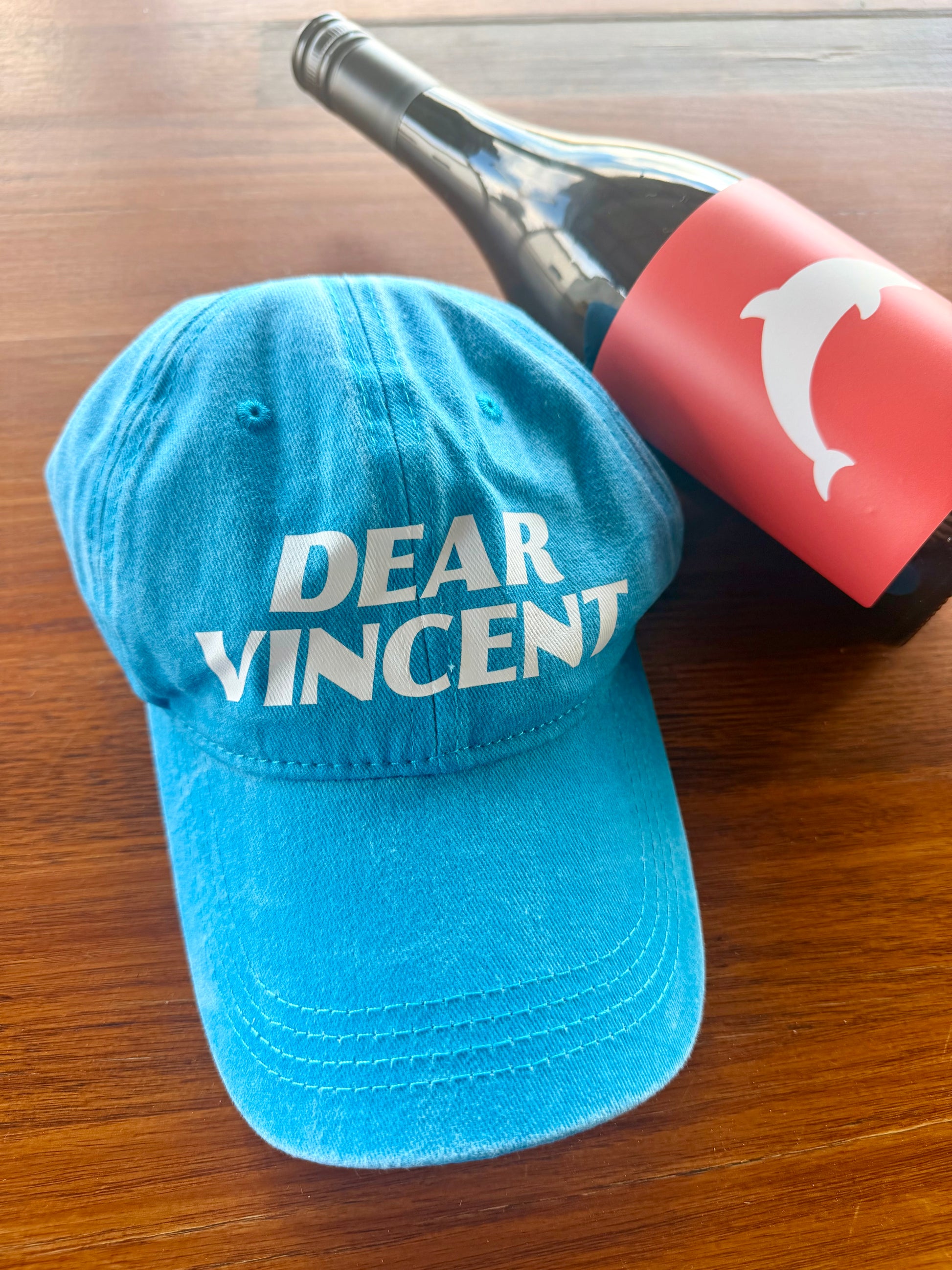 A bright blue denim-style baseball cap with 'DEAR VINCENT' printed in white across the front, placed on a wooden surface next to a wine bottle with a red label.