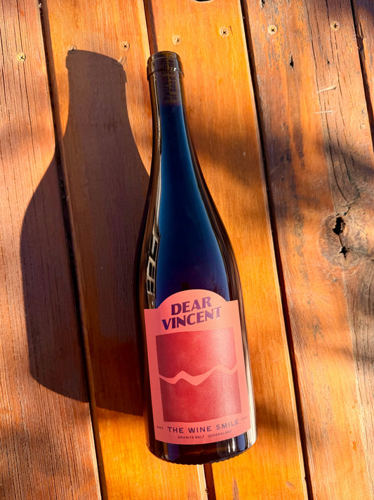 A dark glass wine bottle with a ted label reading 'DEAR VINCENT' and 'THE WINE SMILE' lies on a wooden plank surface in warm sunlight, casting a shadow to the left.